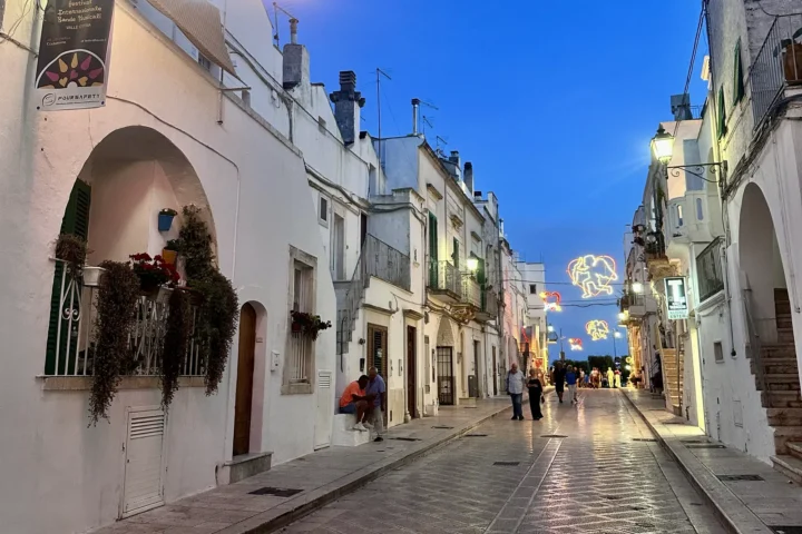 Rua de Cisternino ao anoitecer, com fachadas brancas e iluminação decorativa