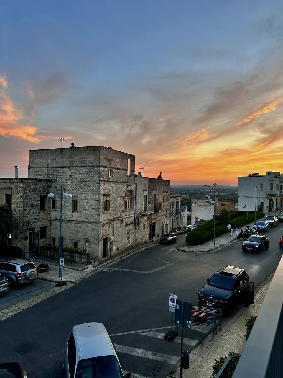 Rua de Cisternino com veículos estacionados e céu colorido ao pôr do sol